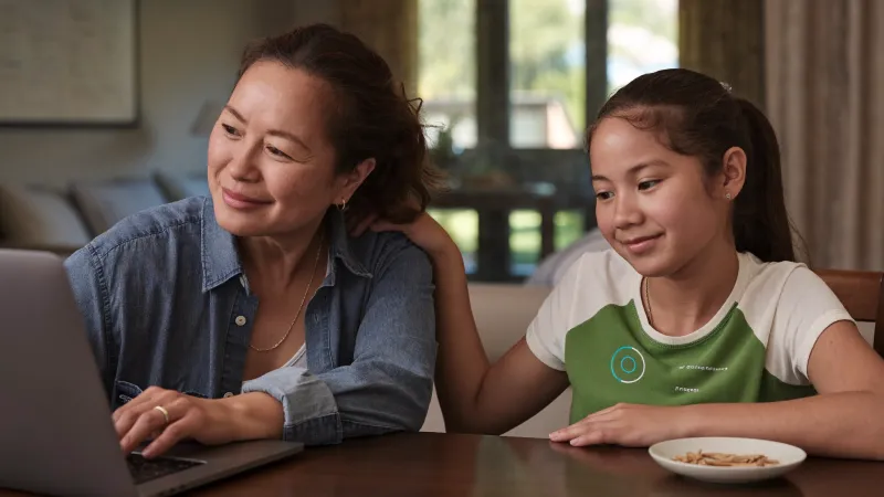 A mother and her teenage daughter sit together at a dining table looking at a laptop, relaxed and proud